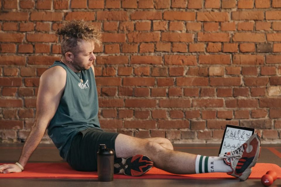 Athletic man sitting and resting on a mat in an urban gym, with a tablet and weights visible.